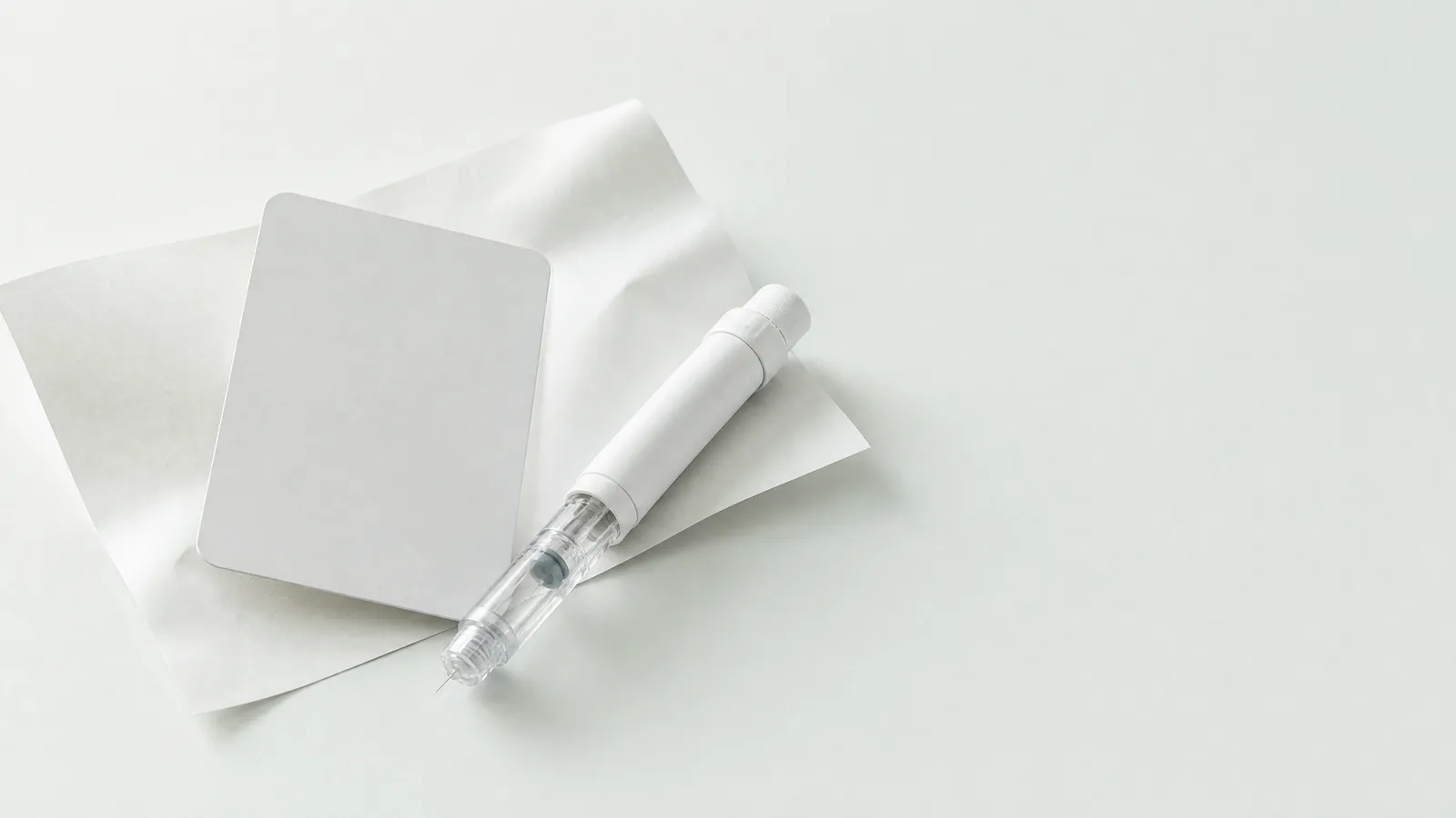 Studio still life: a blank card on a folded sheet of paper next to an unbranded pen injector